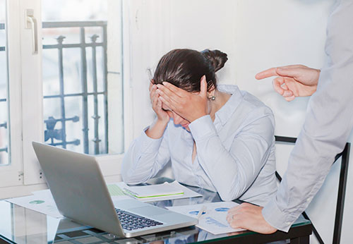 Stressed woman at a desk covering her face while another person points during a tense discussion.